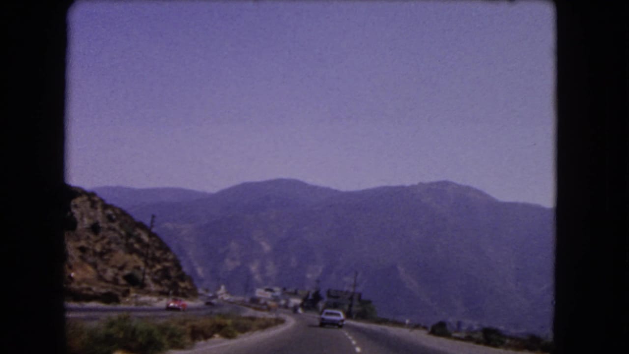 Final 8mm frame of the 1966 California mountain drive, with a car ahead, rocky slope, and hazy peaks.