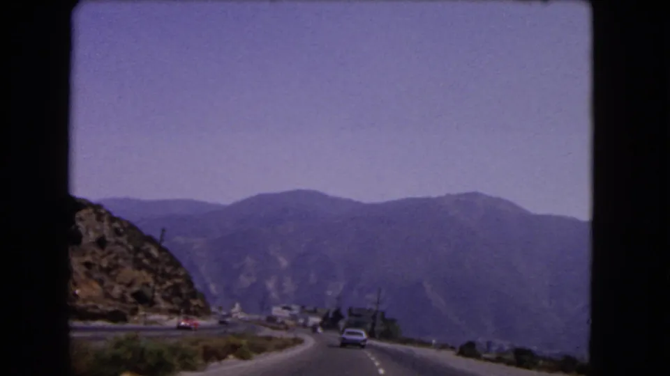 Final 8mm frame of the 1966 California mountain drive, with a car ahead, rocky slope, and hazy peaks.