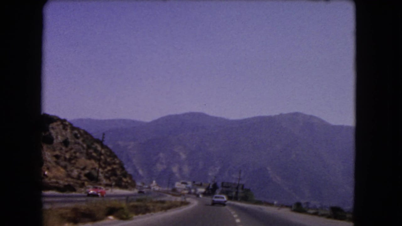 1966 California 8mm frame showing a divided mountain road from a moving car, with period vehicles, dry hillside, and distant ridges.