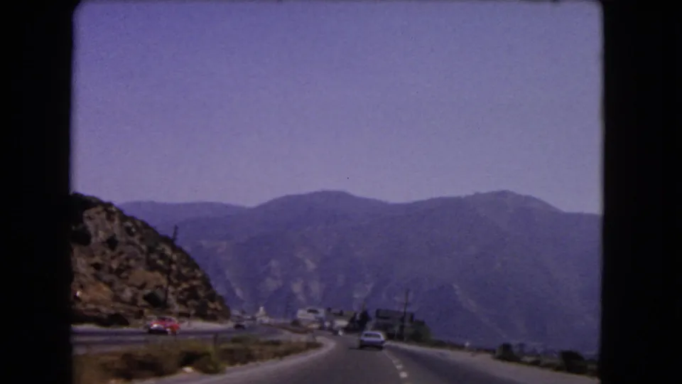 1966 California 8mm frame showing a divided mountain road from a moving car, with period vehicles, dry hillside, and distant ridges.
