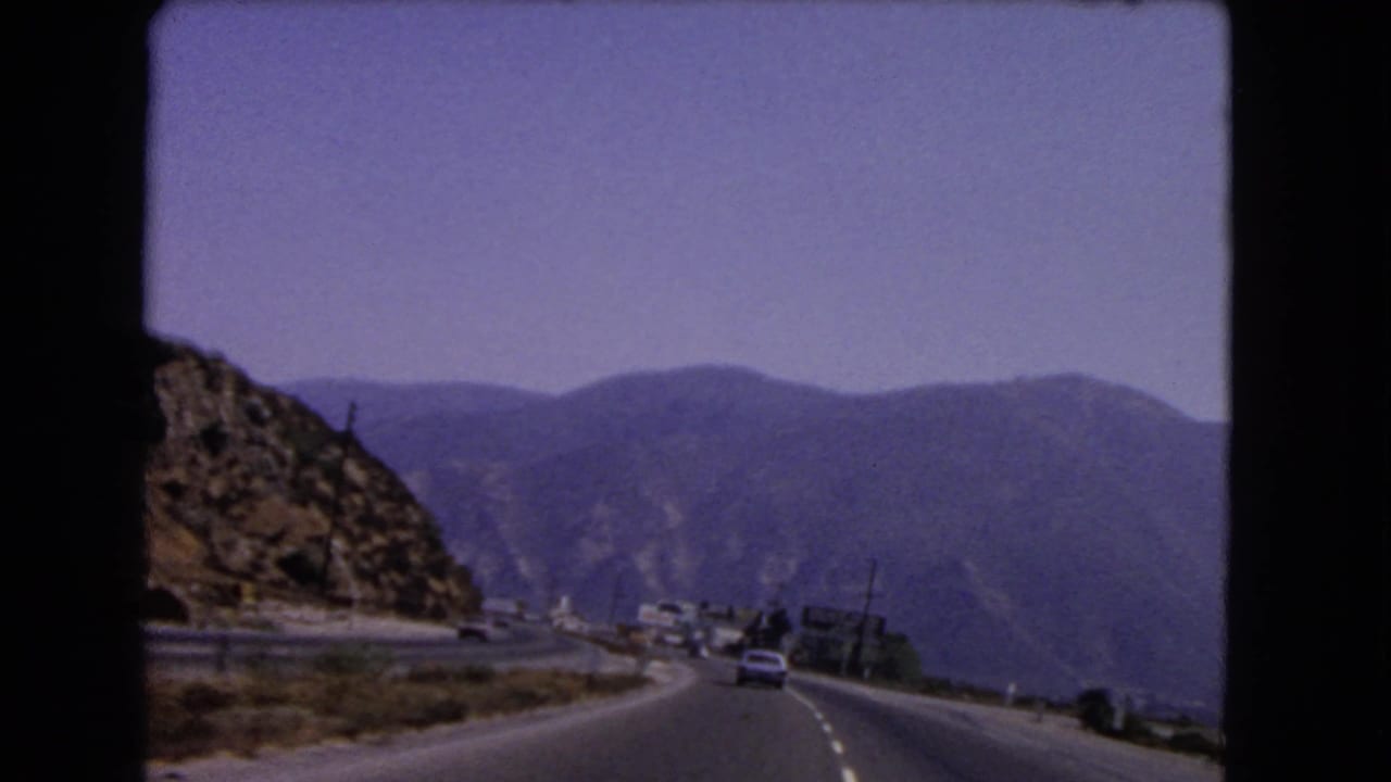 Curving mountain road with cars, roadside signs, and hills in the distance.