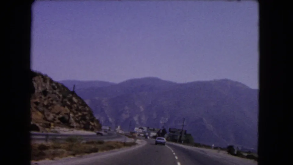 1966 California road footage frame with a curving four-lane mountain highway, visible cars, roadside signs, and hazy ridgelines.