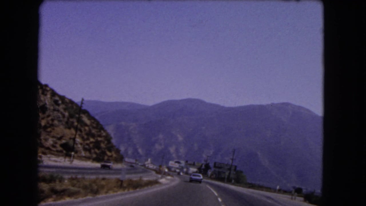 Passenger-view 8mm frame from 1966 California showing a broad mountain road, distant ridges, dry roadside terrain, and cars ahead.