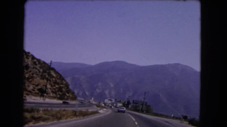 Passenger-view 8mm frame from 1966 California showing a broad mountain road, distant ridges, dry roadside terrain, and cars ahead.