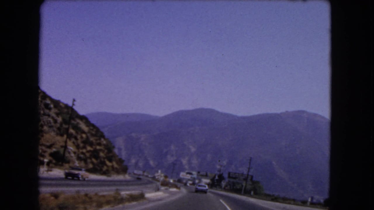 1966 California 8mm frame of a four-lane mountain road curving past a rocky hillside, with traffic and distant mountains visible.