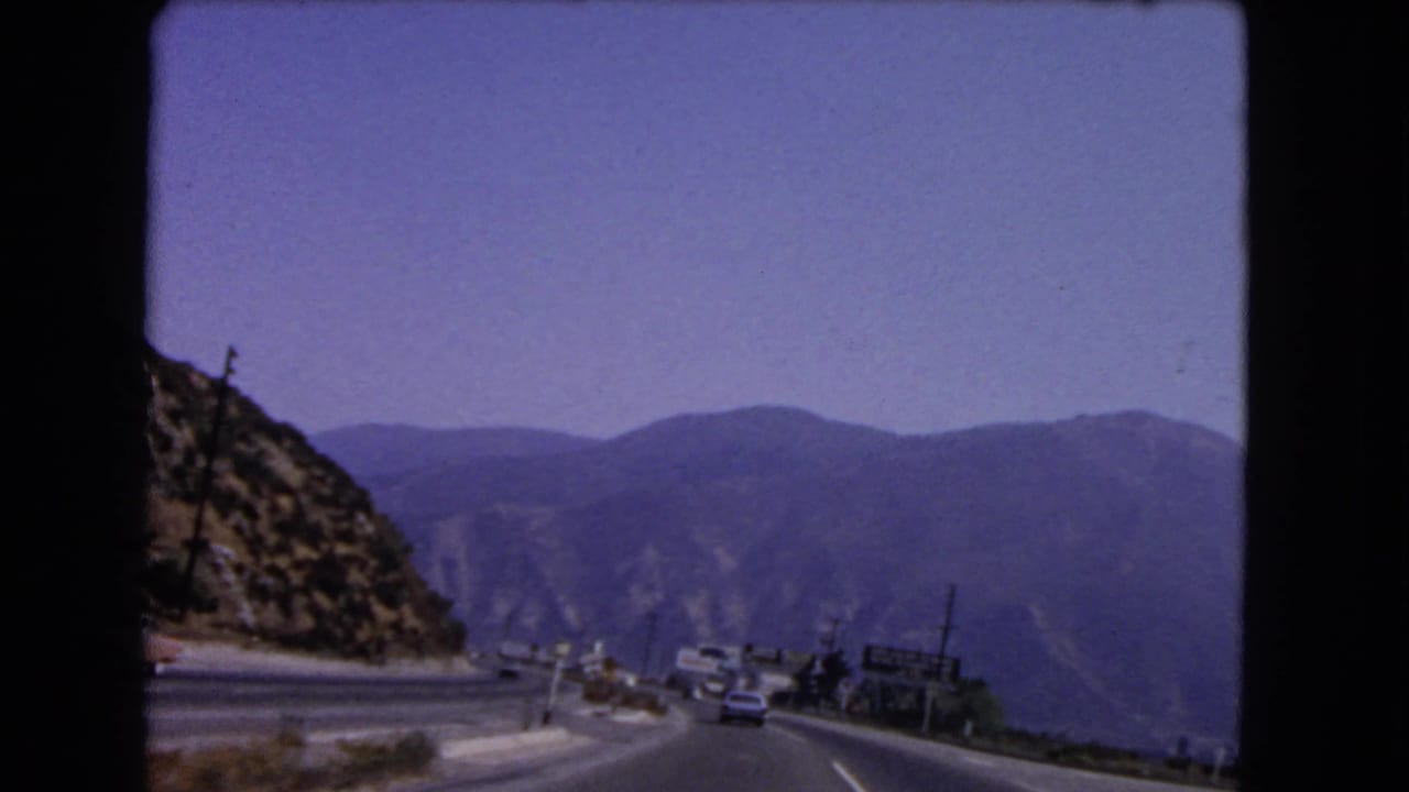 1966 California home-movie view of a mountain highway with cars ahead, roadside signs, utility poles, and hazy ridgelines.