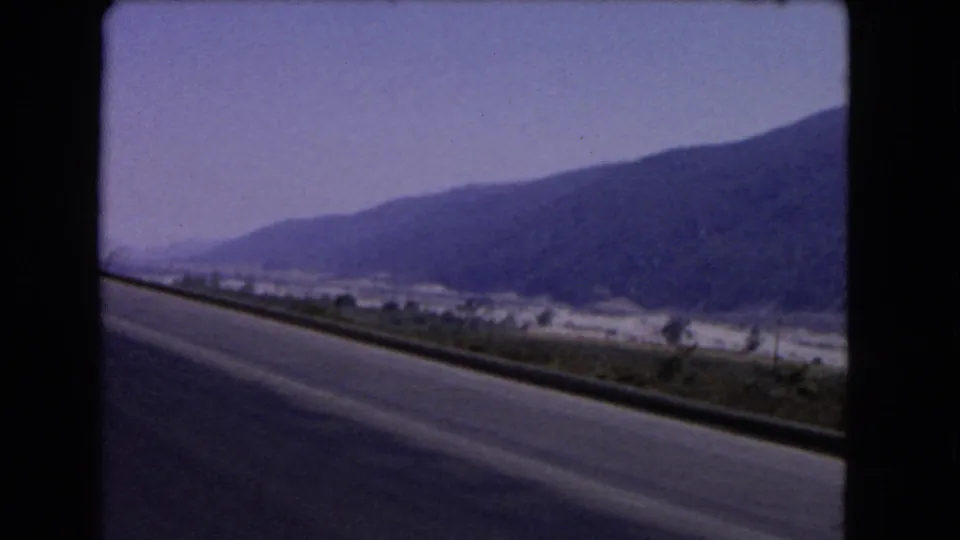 1966 California road-trip frame showing a paved roadway, guardrail, dry valley, and long mountain ridge under a hazy sky.