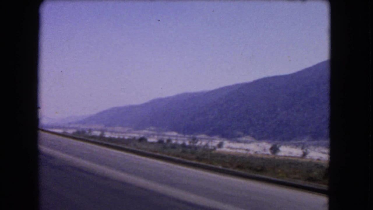1966 California home-movie frame from a moving vehicle, showing a curving roadside guardrail, valley floor, and mountain slope.