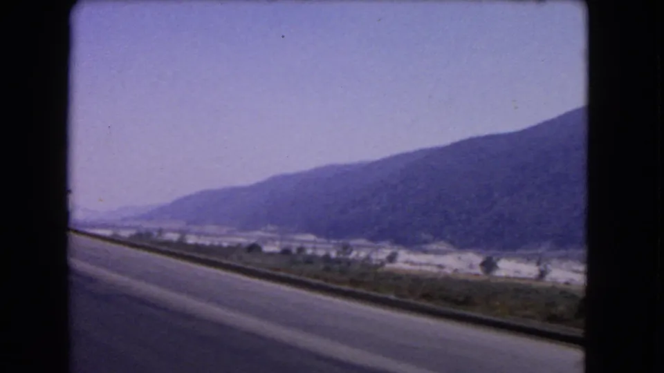 1966 California home-movie frame from a moving vehicle, showing a curving roadside guardrail, valley floor, and mountain slope.