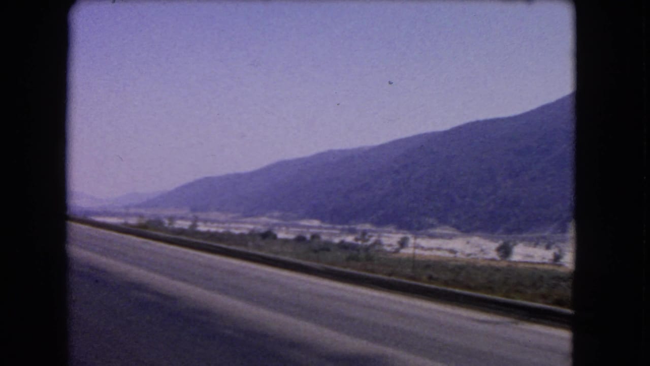1966 California road-trip still showing a paved roadside, guardrail, hazy valley details, and a dark mountain ridge.