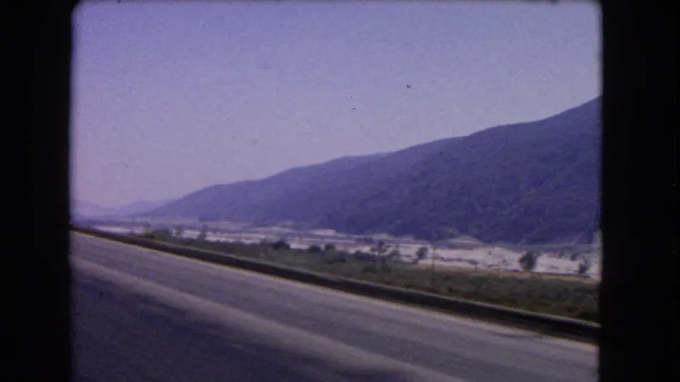 1966 California road-trip still showing a paved roadside, guardrail, hazy valley details, and a dark mountain ridge.
