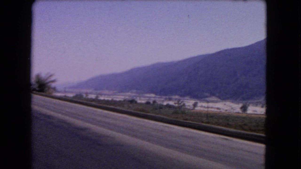 1966 California 8mm road view from a moving car, showing a highway shoulder, valley settlement and mountains in the distance.