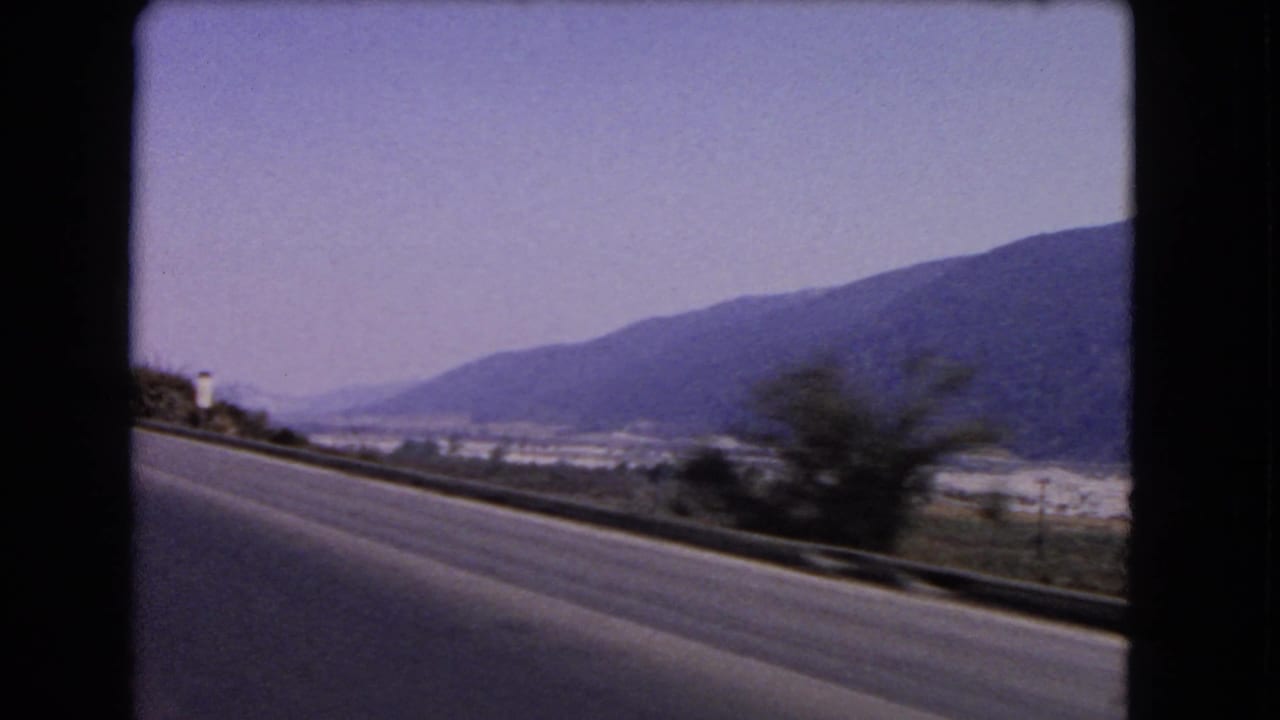 Roadside view of a highway with brush, a distant valley settlement and dark mountains.