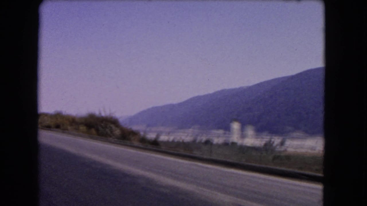 1966 California highway footage showing a roadside curve, white water tower, distant settlement and mountain backdrop from a moving vehicle.