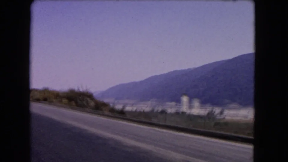 1966 California highway footage showing a roadside curve, white water tower, distant settlement and mountain backdrop from a moving vehicle.