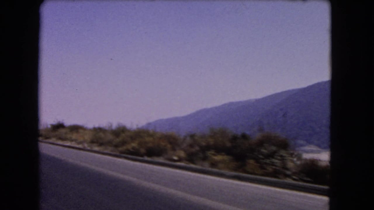 Soft 8mm frame from a 1966 California car trip, showing highway pavement, low roadside vegetation and mountains across the valley.