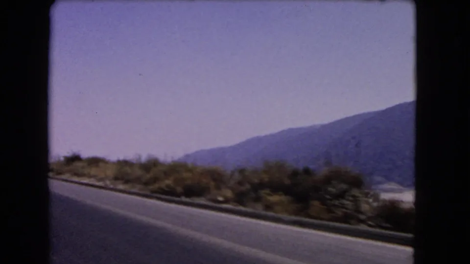 Soft 8mm frame from a 1966 California car trip, showing highway pavement, low roadside vegetation and mountains across the valley.