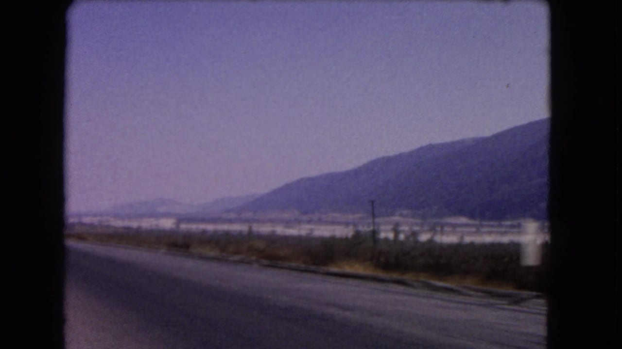 Paved road beside dry roadside plants with mountains and a hazy sky in the distance.