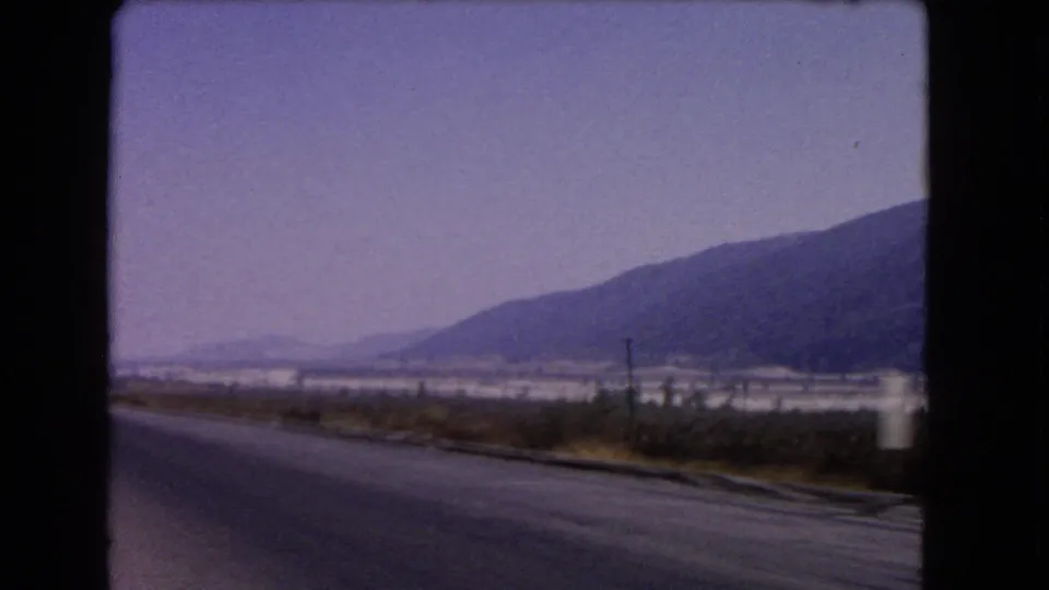 1966 California 8mm frame from a moving vehicle showing a paved road, sparse roadside plants, distant valley, and mountain slopes.