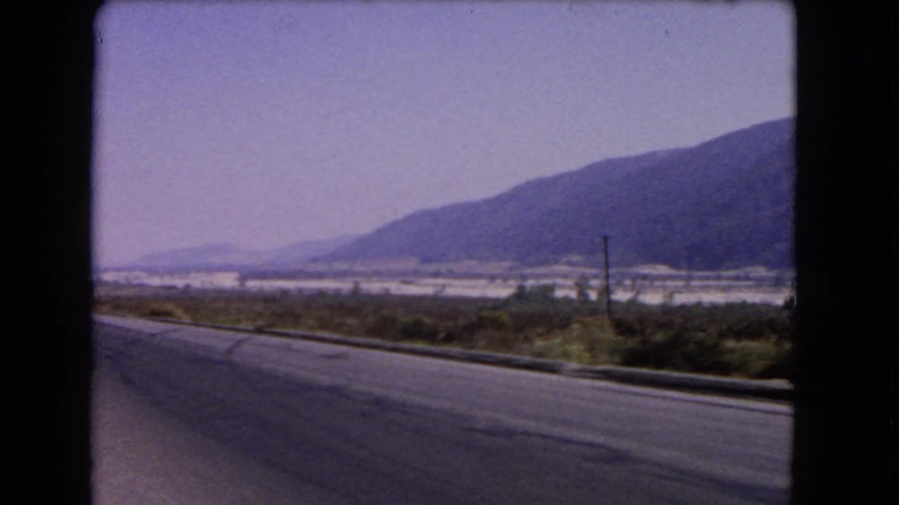 1966 California travel frame showing a road curving past brush and utility poles toward a broad valley and mountain ridge.