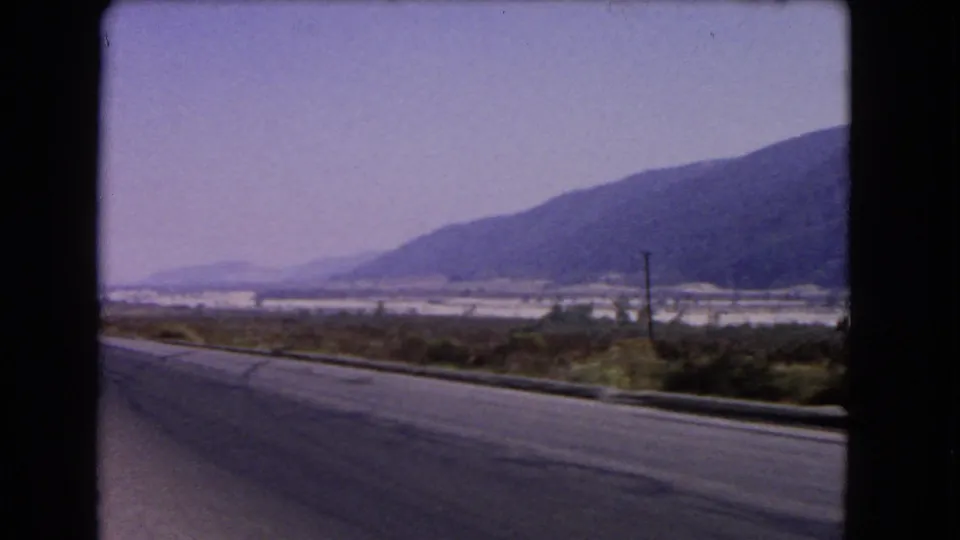 1966 California travel frame showing a road curving past brush and utility poles toward a broad valley and mountain ridge.