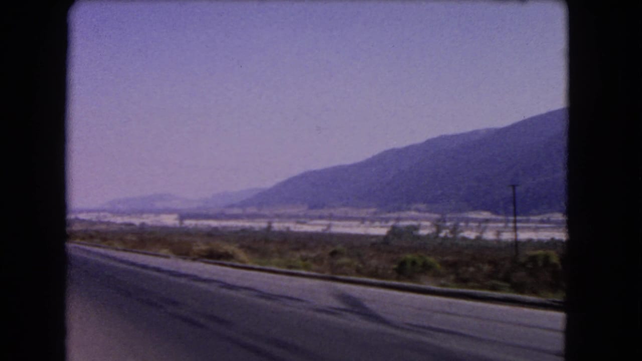 1966 California 8mm frame with a paved road edge, sparse vegetation, a utility pole, light-toned valley area, and mountains beyond.