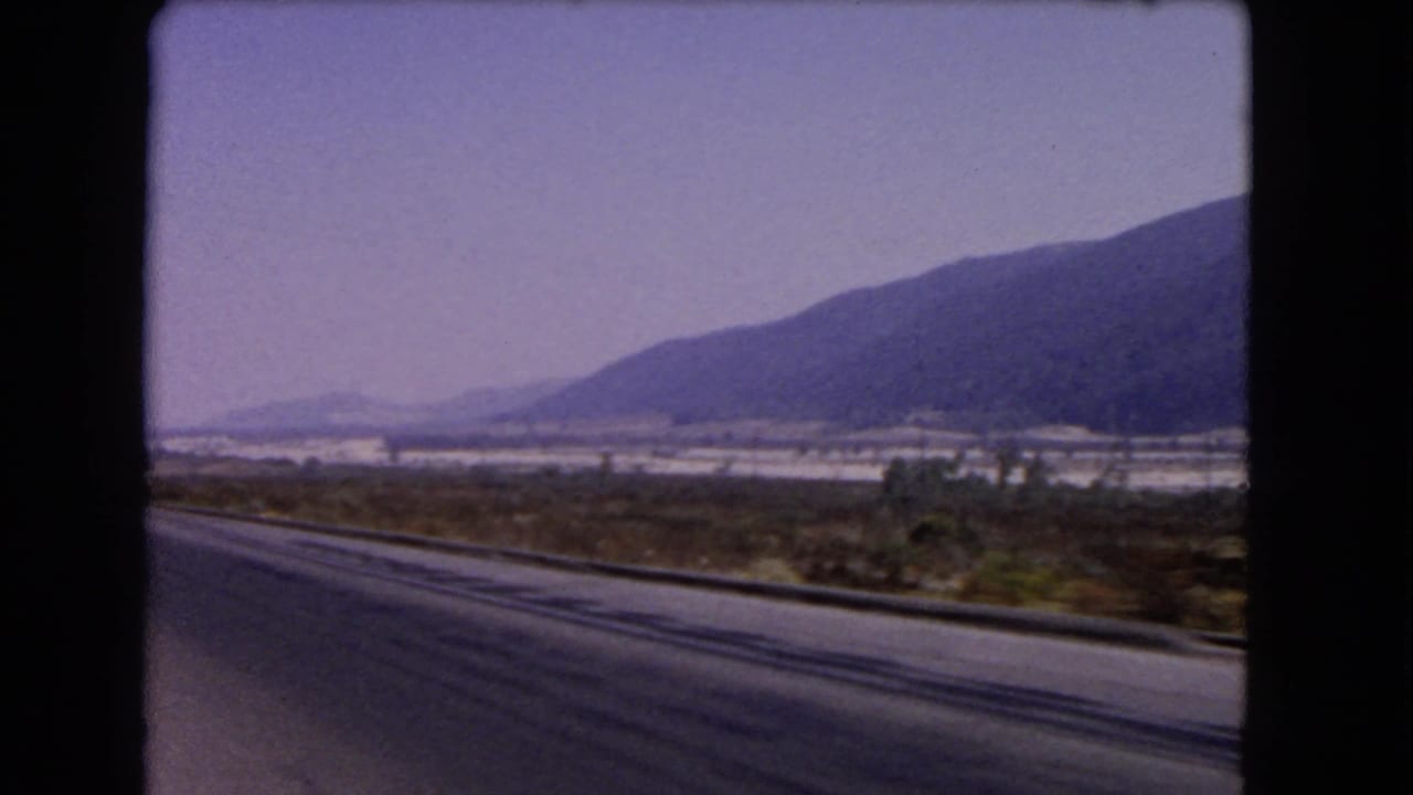1966 California home-movie frame looking across a paved roadside toward brush, a distant valley, and blue-purple mountain slopes.