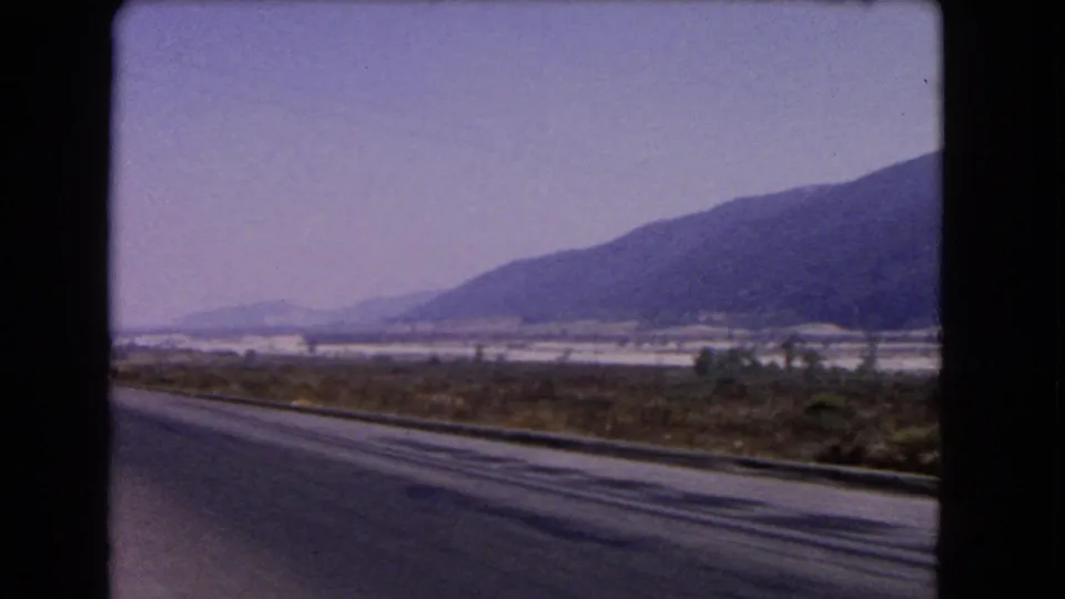 1966 California 8mm road-trip frame showing a mountain slope, open roadside land, and a paved road edge from a moving car.