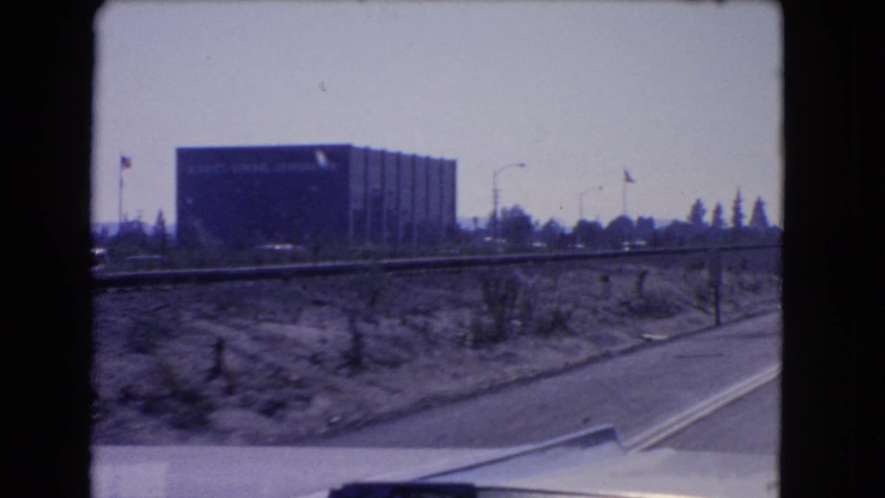 1966 California road-trip frame showing a large Kellogg Company building beyond roadside scrub and a highway edge.