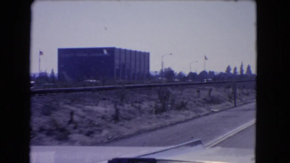 1966 California road-trip frame showing a large Kellogg Company building beyond roadside scrub and a highway edge.