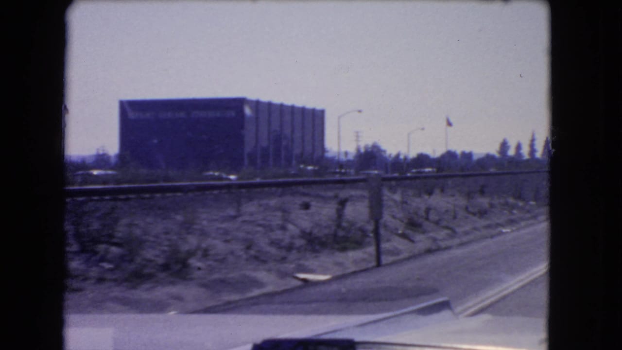 1966 California 8mm frame showing the Kellogg Company building across a road, with scrubland, poles, and car body visible.