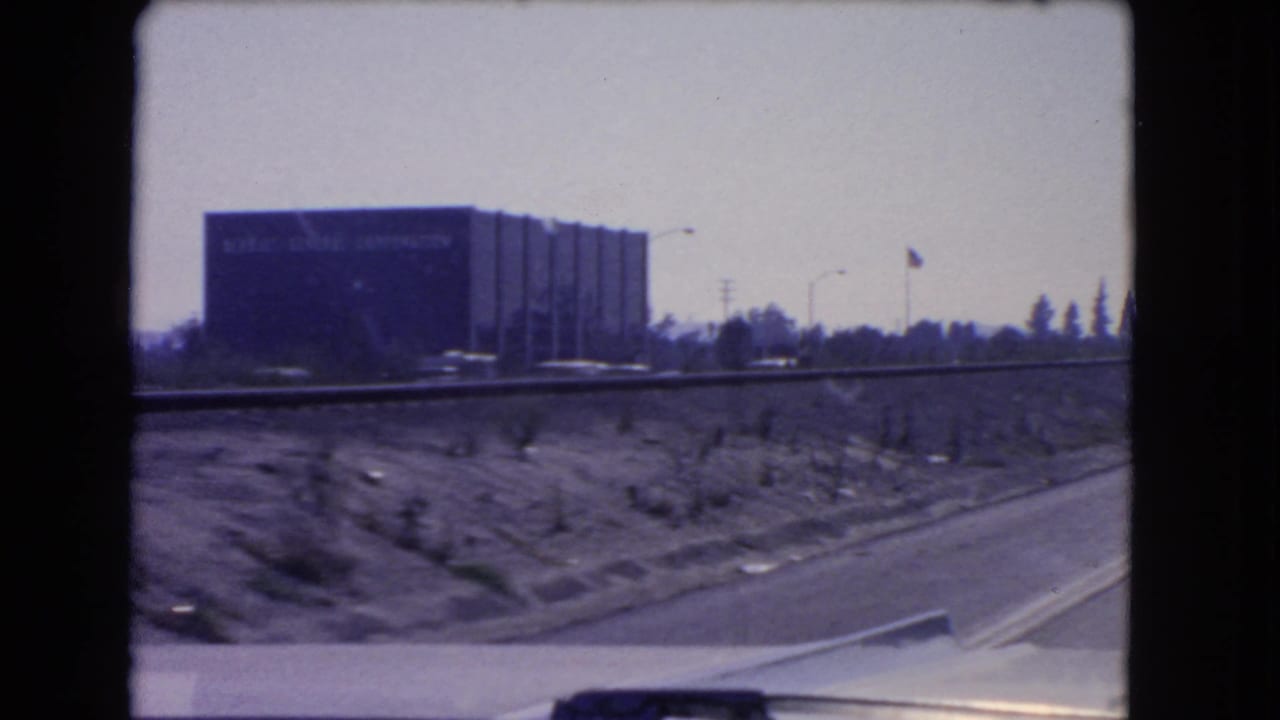 1966 California home-movie frame showing a drive-by view of the Kellogg Company building, open roadside ground, and car hood.