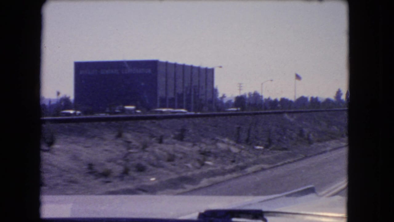 Moving car view of a dark roadside building beyond a freeway embankment.