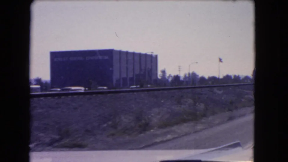 A car-mounted or passenger-view shot passes a dark building beside a California freeway, with traffic and an American flag visible.