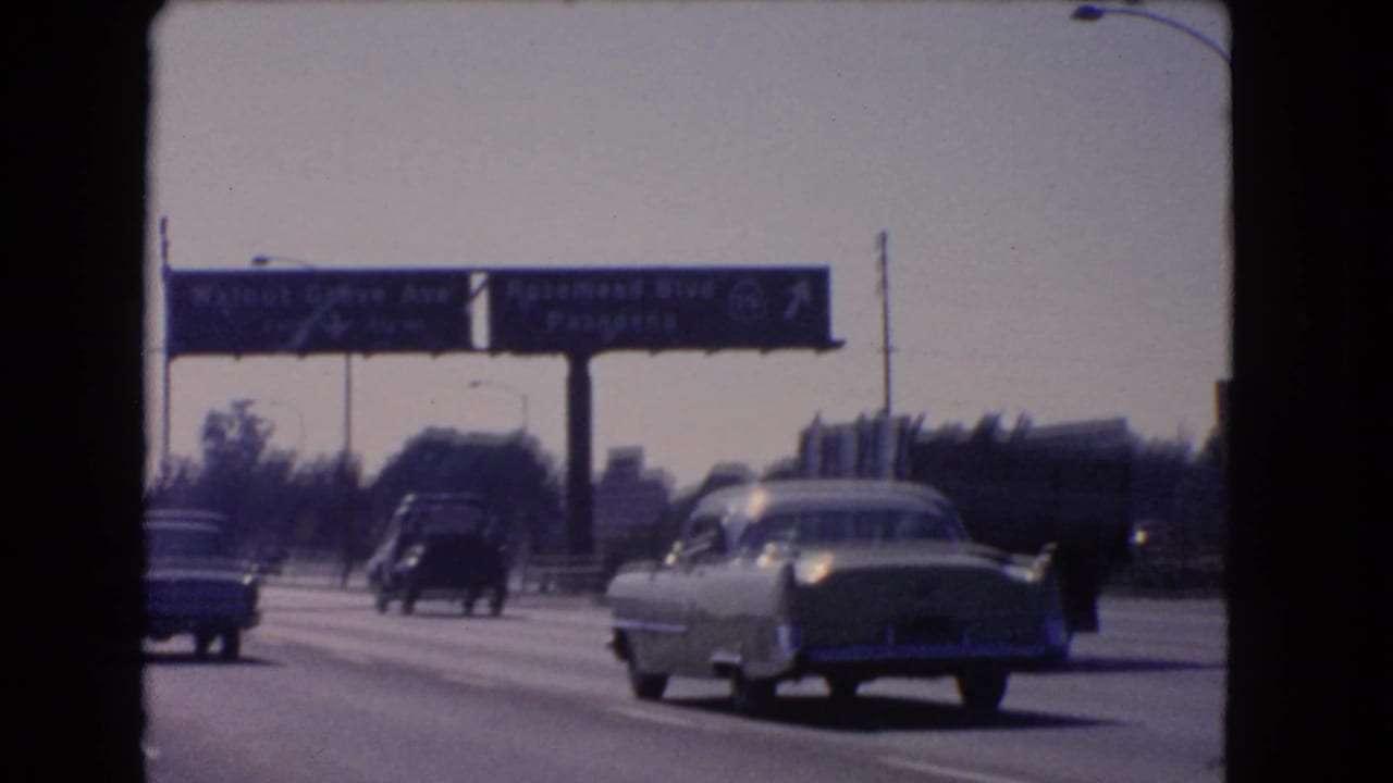 Traffic passes beneath overhead signs on a sunlit multi-lane highway.