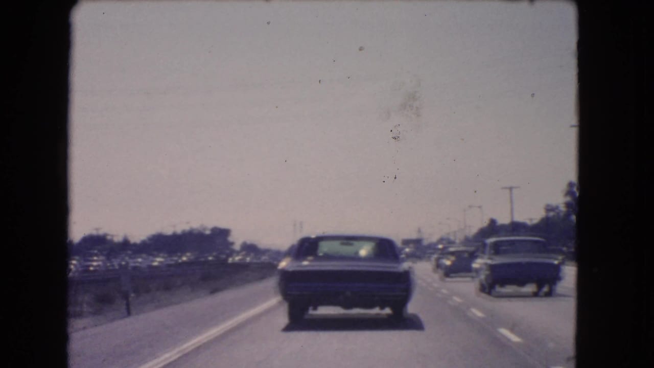 Passenger-shot 8mm view of 1966 California freeway traffic, with cars ahead and road infrastructure in soft focus.