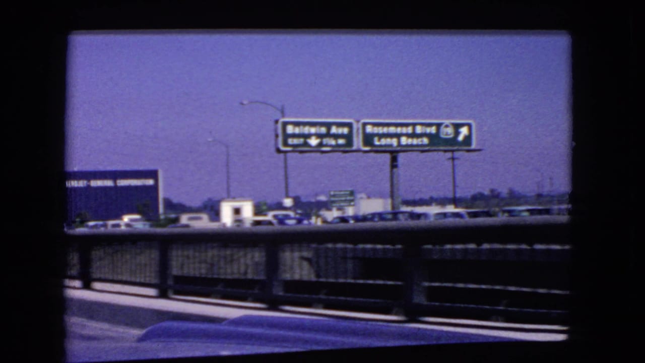 Roadside traffic and freeway direction signs viewed through a dark vehicle window frame.