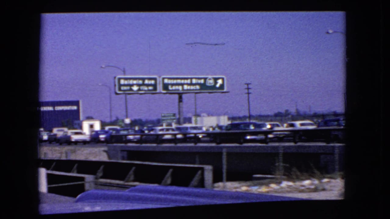 1966 California highway traffic beneath Baldwin Ave and Rosemead Blvd signage, with cars, lamp posts, roadside barriers, and open ground visible.