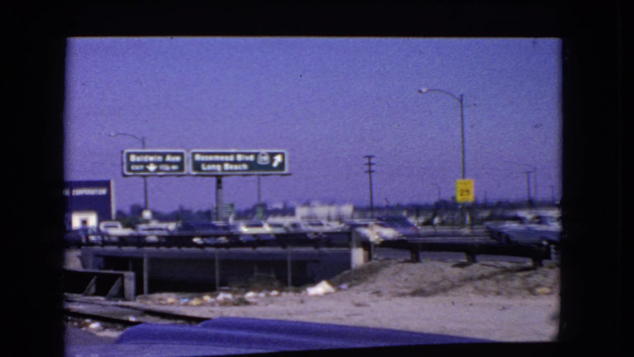 California highway scene from 1966 with Baldwin Ave and Rosemead Blvd signs, distant traffic, lamp posts, a yellow speed sign, and graded roadside earth.