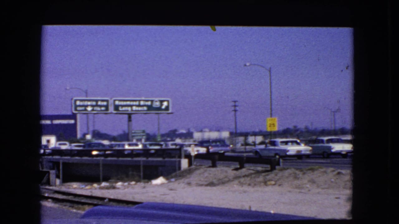 1966 California freeway traffic seen through a windshield, with vintage cars and overhead signs for Baldwin Ave and Rosemead Blvd toward Long Beach.
