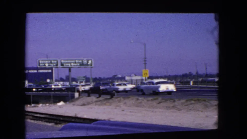 1966 California highway scene with vintage cars, overhead signs toward Long Beach, a yellow speed sign, and the filmed car hood at the bottom edge.
