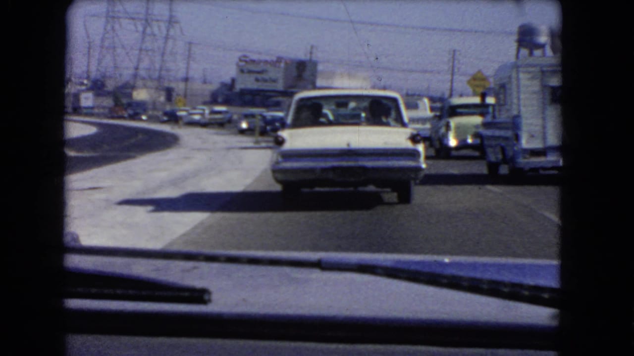 1966 California traffic filmed through a windshield, showing a white sedan ahead, roadside vehicles, power lines, and a distant Smirnoff billboard.
