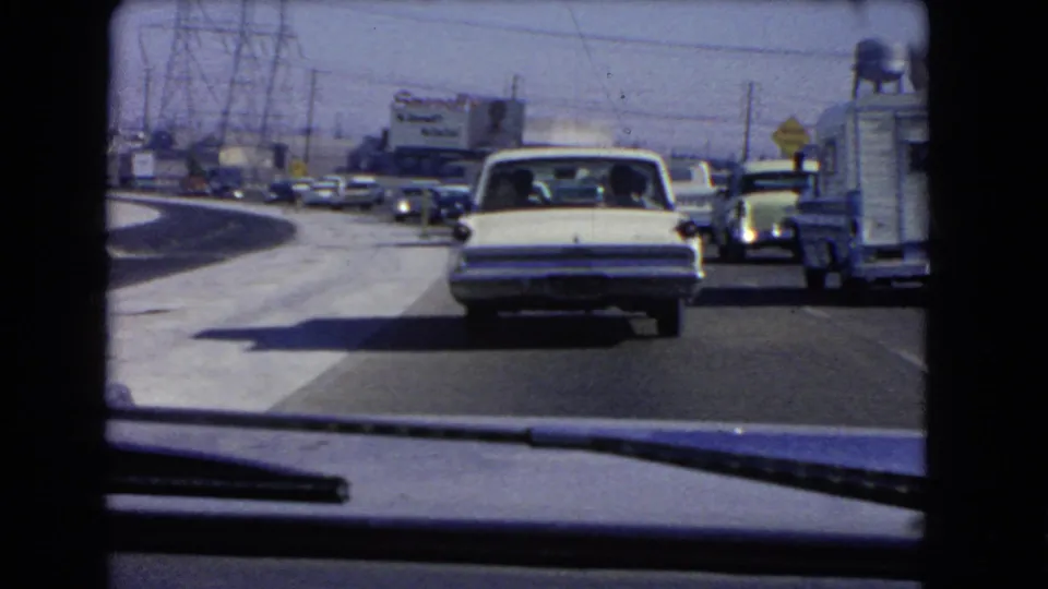 1966 California traffic filmed through a windshield, showing a white sedan ahead, roadside vehicles, power lines, and a distant Smirnoff billboard.
