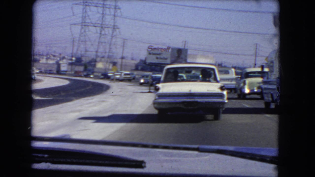 1966 California freeway traffic from inside a car, with a white sedan ahead, adjacent vehicles, power infrastructure, and a Smirnoff billboard in the background.