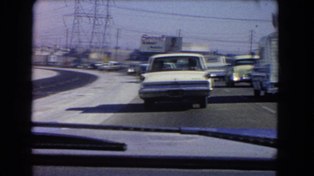 1966 California highway traffic seen from inside a car, with older sedans ahead, power lines, and a dashboard edge in view.