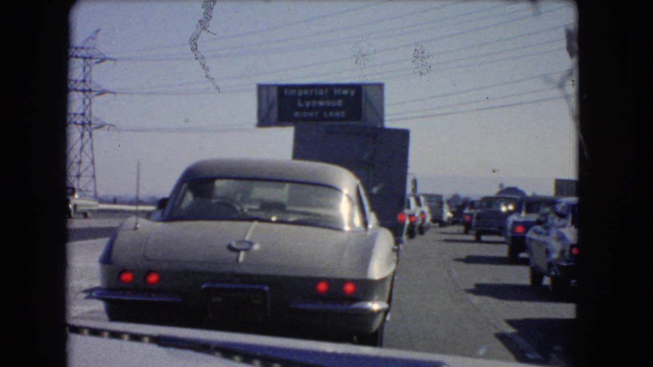 1966 California traffic jam with a silver Corvette ahead and an overhead sign reading Imperial Hwy and Lynwood right lane.