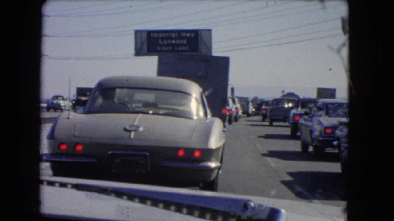 Rear view of a silver Corvette in a 1966 California highway jam, with cars filling adjacent lanes and an Imperial Hwy sign above.