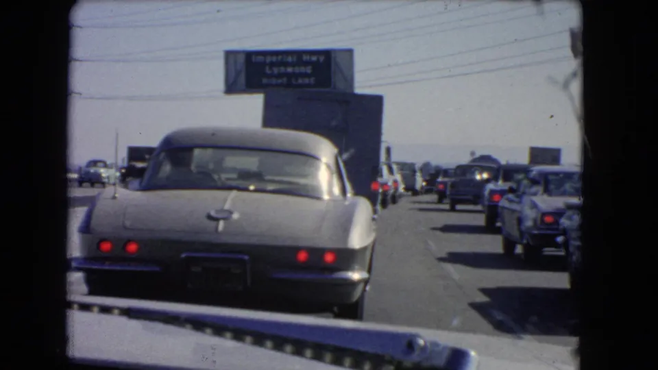Rear view of a silver Corvette in a 1966 California highway jam, with cars filling adjacent lanes and an Imperial Hwy sign above.