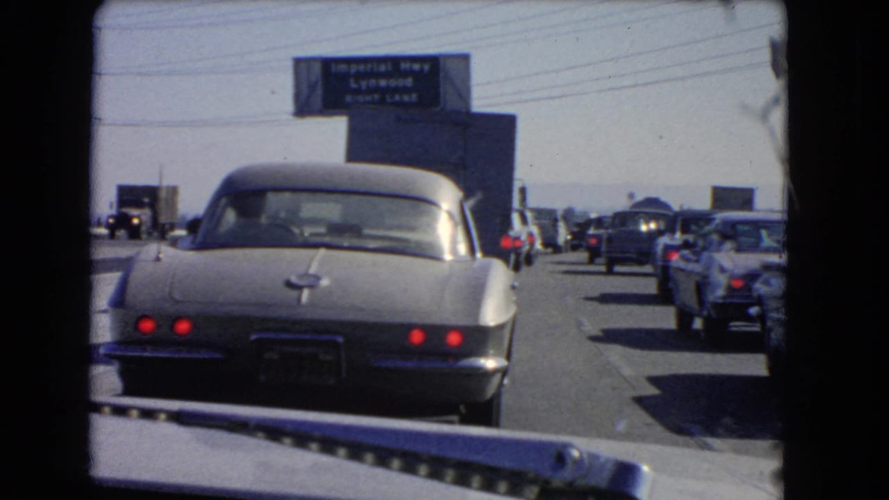 1966 California freeway congestion viewed through a windshield, with a silver Corvette ahead and multiple lanes of older cars near Lynwood signage.
