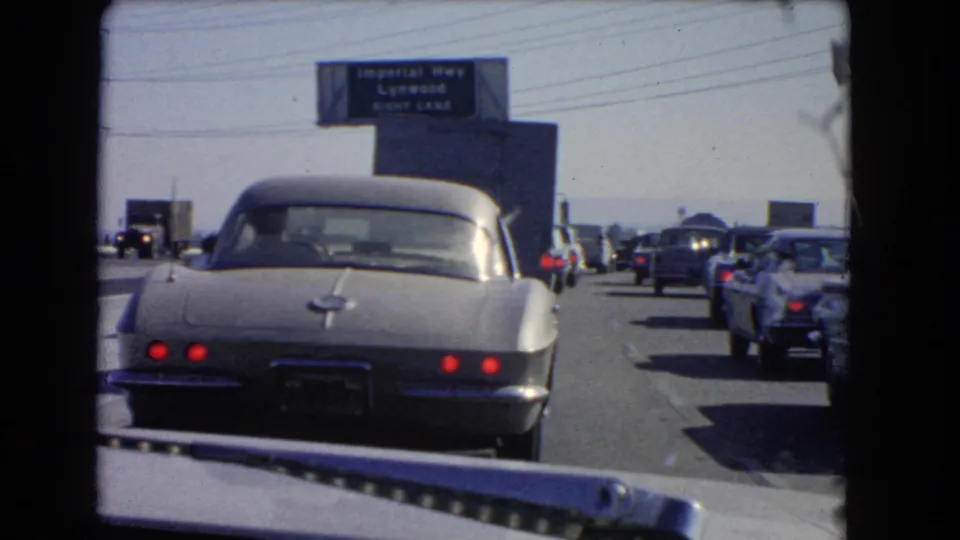 1966 California freeway congestion viewed through a windshield, with a silver Corvette ahead and multiple lanes of older cars near Lynwood signage.